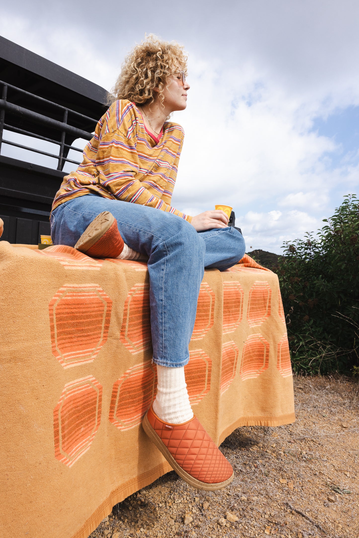 Lifestyle photo of a women sitting down looking in the distance while wearing the Malibu sandals westward ballistic clog in rust 