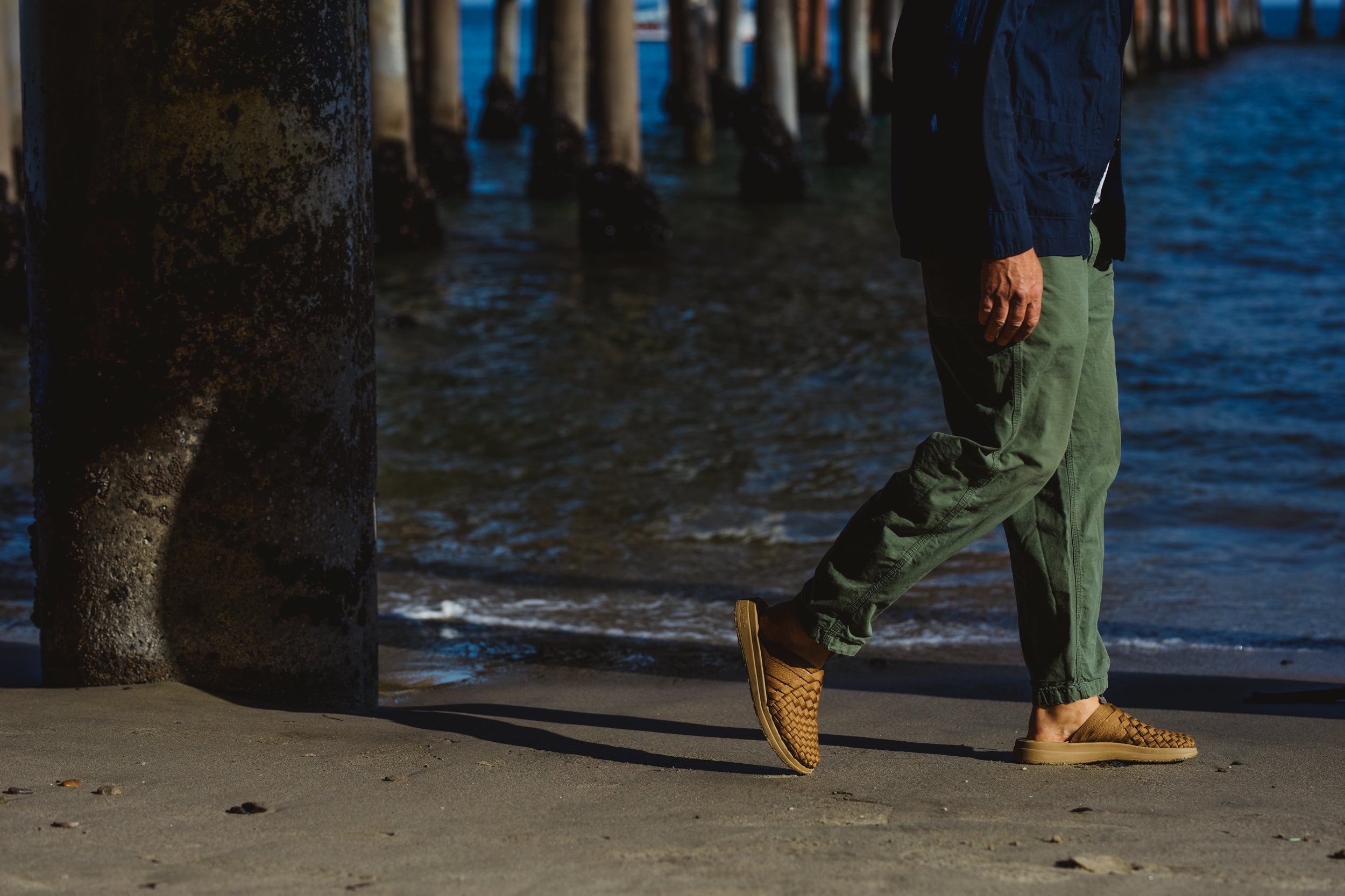 malibu sandals lifestyle photo of a person wearing the coyote nylon colony sandals with green pants and blue over shirt on the beach with the pier background