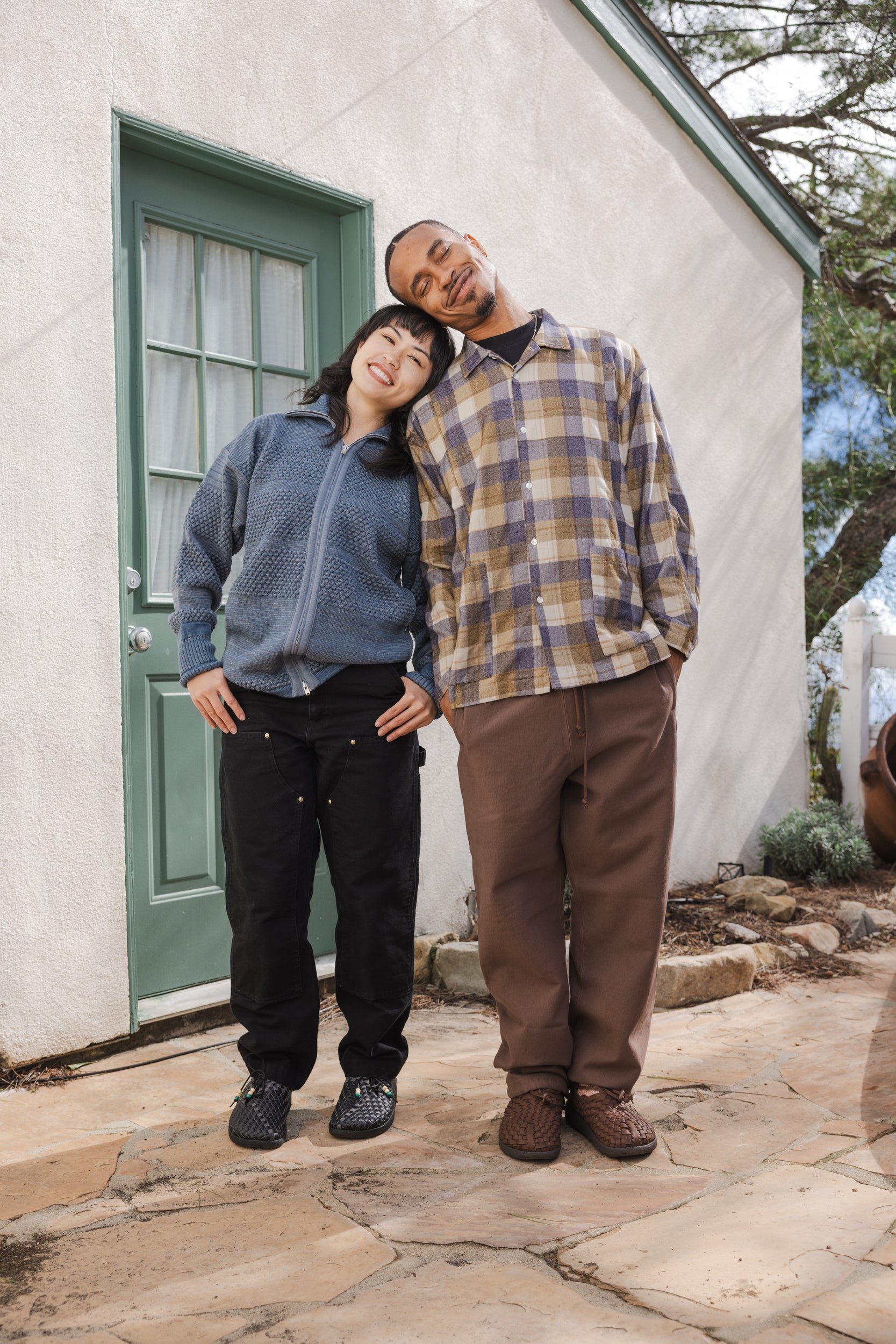 Lifestyle photo of an Asian female and black male both standing and leaning on each other. Female is a blue sweater, black pants and black Malibu sandals Black Matador. Male is wearing plaid over shirt brown pants and Malibu sandals bison colored Latigo moc. 