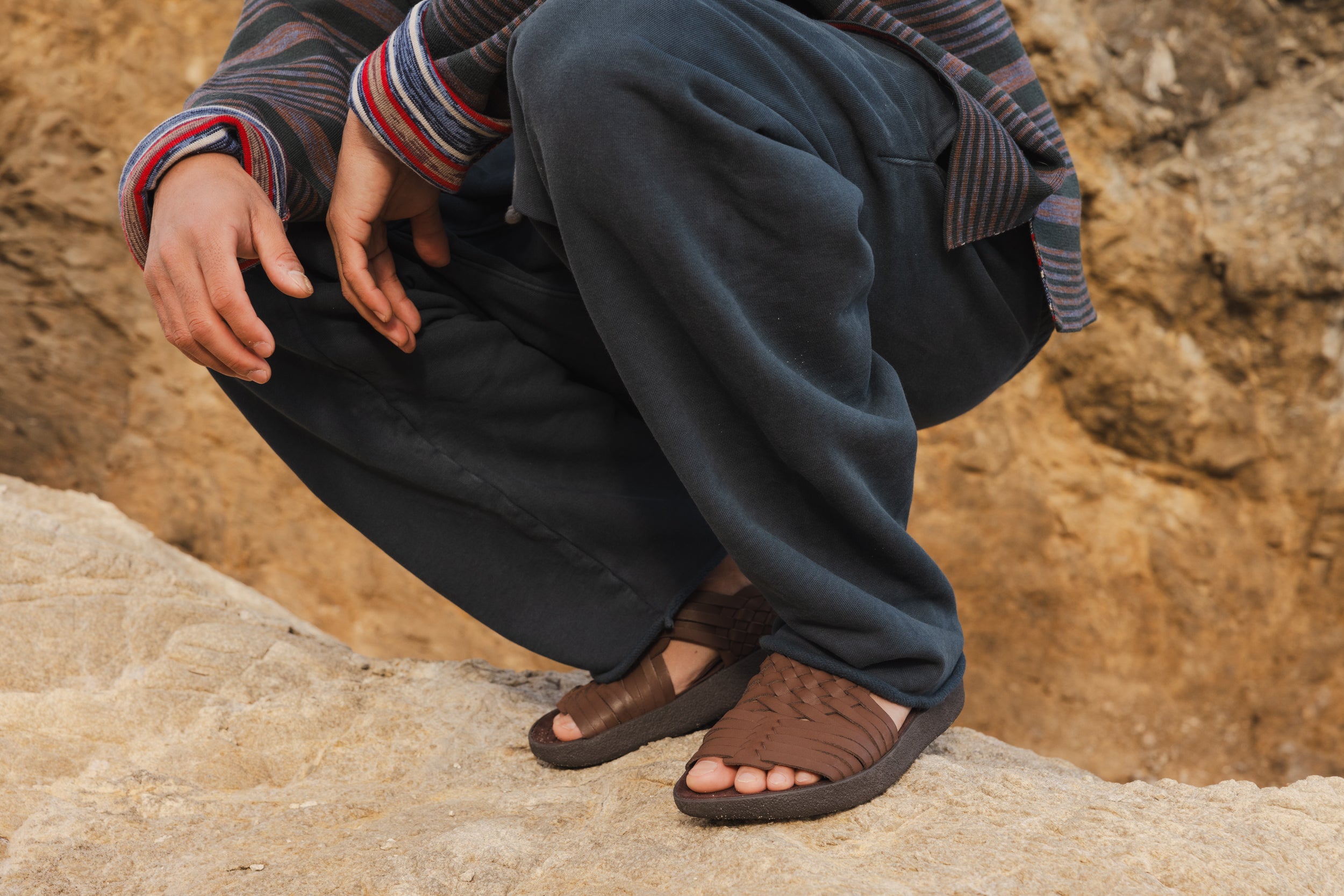 Person squatting on a beach rock wearing Malibu sandals vegan leather canyon in Bison colorway with blue sweaters and a stripped sweater.