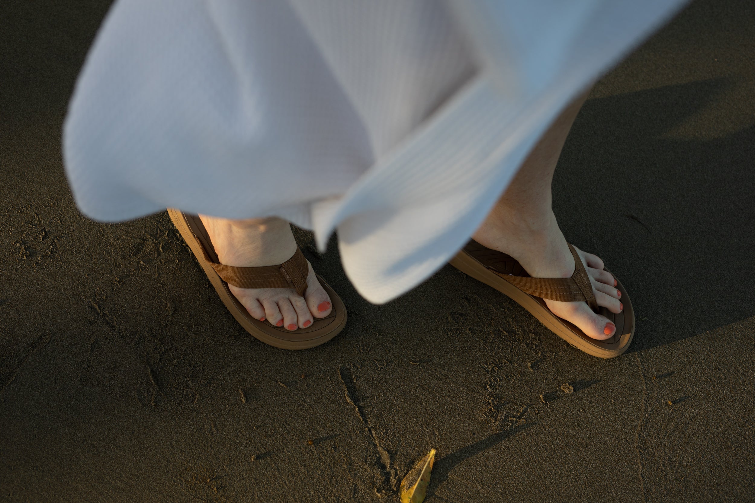 lifestyle macro phot of malibu sandals surfrider style in coyote color being worn with a white dress on the wet soft beach sand